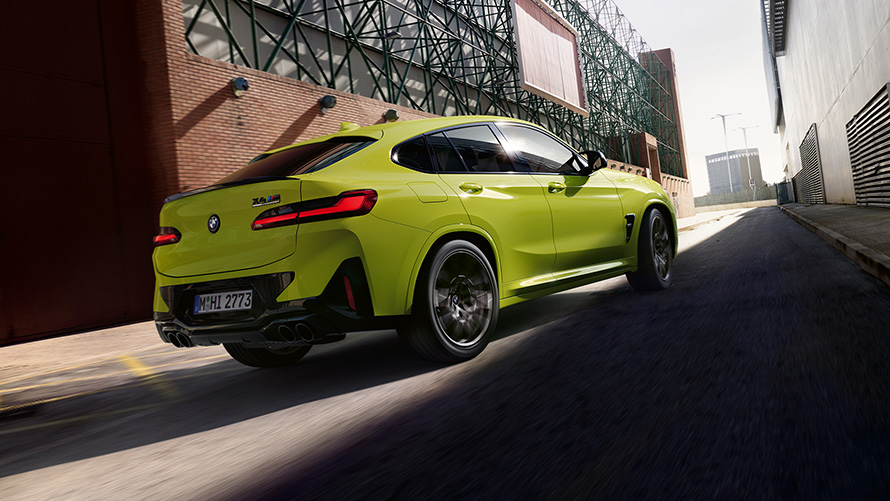 BMW X4 M Competition F98 LCI Facelift 2021 Sao Paulo Yellow Vista posteriore a tre quarti in movimento tra capannoni